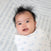 A baby with spiky black hair is wrapped in a HALO Swaddle 100% Cotton TOG 1.5, showcasing a white blanket with blue patterns. The adjustable wings cradle the baby, whose wide eyes and small smile brighten the soft, light-colored textured background.