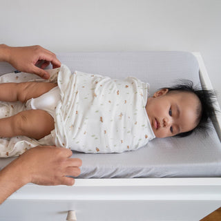 A baby is snuggled into a HALO Swaddle 100% Cotton TOG 1.5 on a changing table, wrapped in a white blanket with animal prints. Featuring adjustable wings, the swaddle ensures healthy hip development as gentle hands secure it around the baby on a soft gray surface.