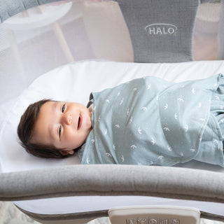 A baby rests in a gray bassinet, swaddled comfortably in HALOs Swaddle 100% Cotton TOG 1.5, light blue with white patterns. The infant smiles at the camera, showcasing dark hair and a cheerful face. The HALO logo signifies superior quality and supports healthy hip growth.