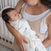 In a softly lit room, a woman cradles her sleeping baby snugly wrapped in a HALO Swaddle 100% Cotton TOG 1.5 with adjustable wings and a double zipper. Content with a pacifier, the baby rests peacefully in her arms next to the crib.