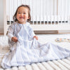 A toddler wearing a white and blue striped HALO Wearable Blanket 100% Cotton TOG 0.5 sits on a white carpet in a bright room, smiling. A crib is in the background, and a toy is visible on the floor, providing cozy and safe sleep.
