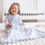 A toddler wearing a white and blue striped HALO Wearable Blanket 100% Cotton TOG 0.5 sits on a white carpet in a bright room, smiling. A crib is in the background, and a toy is visible on the floor, providing cozy and safe sleep.