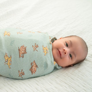 A baby lies on a white sheet, gently gazing up while wrapped in a Disney Lion King 100% Cotton Swaddle TOG 1.5 by HALO, featuring cartoon animals.