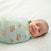 A baby lies on a white sheet, gently gazing up while wrapped in a Disney Lion King 100% Cotton Swaddle TOG 1.5 by HALO, featuring cartoon animals.