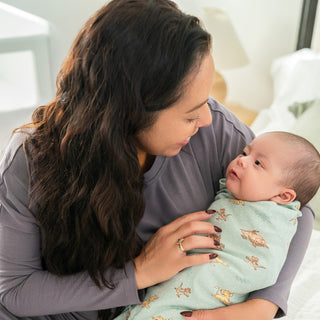 A woman with long dark hair gently holds and smiles at an infant wrapped in a Disney™ Lion King 100% Cotton Swaddle TOG 1.5 by HALO. They are indoors, near a window with soft lighting.