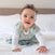 A curly-haired baby with wide eyes crawls on a white bed, wrapped in a HALO Disney™ Lion King 100% Cotton Wearable Blanket TOG 0.5 adorned with playful prints. The light blue outfit contrasts charmingly against the softly blurred neutral-toned headboard, highlighting baby sleep essentials.
