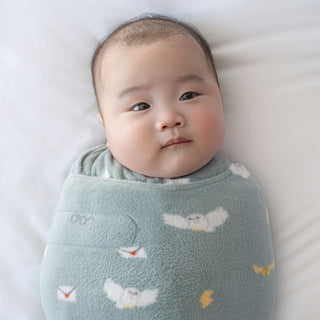 A baby lies calmly on a white surface, wrapped in a soft, light blue HALO Harry Potter™ Swaddle Micro-Fleece TOG 3.0 with white bird patterns, gazing gently to the side.