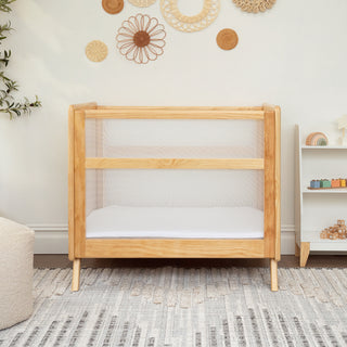 A HALO Mini Breathable Mesh Crib sits by a wall adorned with woven hangings. To the right, a small white shelf holds colorful toys. A textured rug and plush chair complete the cozy nurserys compact footprint.
