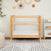 A HALO Mini Breathable Mesh Crib sits by a wall adorned with woven hangings. To the right, a small white shelf holds colorful toys. A textured rug and plush chair complete the cozy nurserys compact footprint.