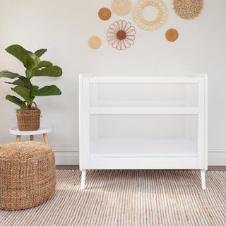 A minimalist nursery features the HALO Mini Breathable Mesh Crib in white, complemented by a white side table with a potted plant and a round woven ottoman. Circular woven wall decorations enhance the rooms look, while a beige striped rug adds warmth to the floor.