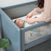 In a HALO Mini Breathable Mesh Crib, a dressed-up baby gazes up as someone adjusts the blanket. The cribs airy mesh sides highlight its compact design, with a plant visible in the corner.