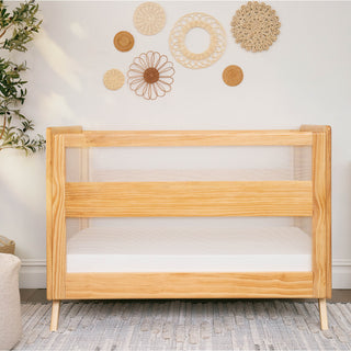 The HALO 3-in-1 Convertible Full Size Breathable Mesh Crib with GREENGUARD GOLD certification and white mesh sides sits on a light blue patterned rug, with woven wall decor and a potted plant adding cozy, modern style to the nursery.