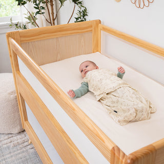 A baby lies on its back in a HALO 3-in-1 Convertible Full Size Breathable Mesh Crib made from New Zealand Pinewood, wearing a nature print sleep sack and green long-sleeve shirt in a softly lit nursery with plants and neutral decor.