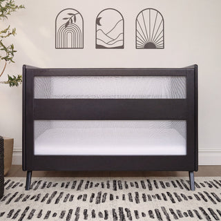 A HALO 3-in-1 Convertible Full Size Breathable Mesh Crib in black stands on a patterned rug, with three minimalistic black wall art pieces—abstract sun, mountain, and rainbow—above. A leafy plant sits to the left.