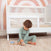 A young child in green pajamas plays with a wooden toy train on a striped rug in front of the HALO 3-in-1 Convertible Full Size Breathable Mesh Crib, now set up as a toddler bed. A painted rainbow arch decorates the wall in the background.