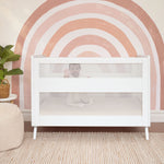 A baby smiles and reaches up while sitting in the HALO 3-in-1 Convertible Full Size Breathable Mesh Crib, set against a wall with a bold pink and orange rainbow mural. A plant, woven basket, and striped rug complete the cozy nursery scene.