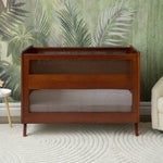 The HALO 3-in-1 Convertible Full Size Breathable Mesh Crib with JPMA certification stands on a white textured rug before leafy green wallpaper, flanked by a white side table and beige armchair.