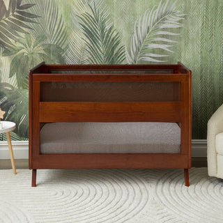 The HALO 3-in-1 Convertible Full Size Breathable Mesh Crib with JPMA certification stands on a white textured rug before leafy green wallpaper, flanked by a white side table and beige armchair.