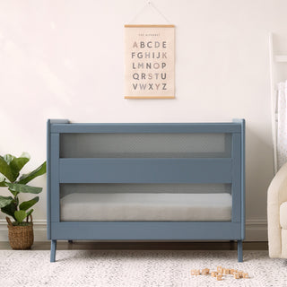 A modern blue HALO 3-in-1 Convertible Full Size Breathable Mesh Crib sits against a light wall below an alphabet poster, with a potted plant to the left, a beige armchair to the right, and wooden blocks on a gray patterned rug.