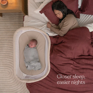 A woman sleeps on burgundy bedding beside a baby in the HALO BassiNest® Lite Portable Bassinet featuring breathable mesh walls. Closer sleep, easier nights decorates the bedspread. The room has a light carpet and wooden nightstand for comfort at adjustable heights.