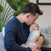 A man cradles a baby wrapped in the HALO SuperSoft Bamboo Swaddle TOG 2.0, gently kissing the baby's head as they sit on a chair with green plants in the background.