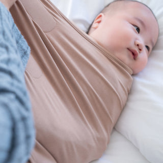 A baby, calm and relaxed, lies on a white bed wrapped in the light brown 3-in-1 swaddle from the HALO Nest-to-Crib Bundle. A soft blue blanket and part of the breathable crib mattress are visible in the foreground.