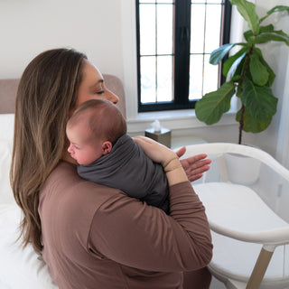 A woman with long brown hair holds a swaddled newborn baby on her shoulder in a cozy, sunlit room with a large window and a green potted plant in the background.