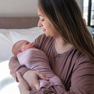 A woman with long brown hair, wearing a mauve top, cradles a swaddled newborn baby in a softly lit bedroom. Both appear calm and content, sharing a tender moment together.