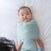 A baby wrapped in a light blue HALO Swaddle SuperSoft Bamboo TOG 2.0 lies on a white bed, looking up as an adult’s hands gently support their sides.