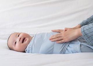 A smiling baby is lying on a white bed, wrapped in a light blue swaddle. An adults hands are gently touching the babys chest, as if soothing or comforting them.