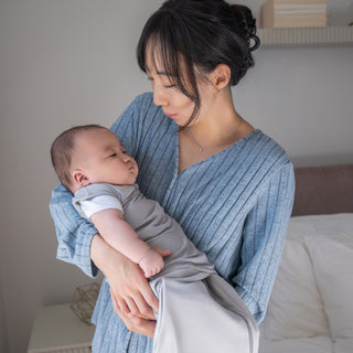 A woman in a blue ribbed sweater gently holds a baby wrapped in a gray blanket, looking down at the baby with a caring expression in a softly lit indoor setting.
