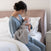 A woman in blue pajamas sits on a bed, gently cradling her baby wrapped in the HALO SuperSoft Bamboo Swaddle TOG 2.0. Both look calm and cozy in a softly lit bedroom.