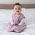 A smiling baby with curly hair sits on a bed, wearing the HALO SuperSoft Bamboo Wearable Blanket TOG 1.5 in light pink over a white long-sleeve shirt, with a white comforter and light brown headboard in the background.