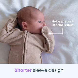 A baby in a beige onesie lies on a white blanket with one arm bent up, cozily wrapped in our hip-healthy HALO SwaddleStretch™ Newborn Swaddle TOG 0.5. Text reads Helps prevent startle reflex. A caption below says, Shorter sleeve design.