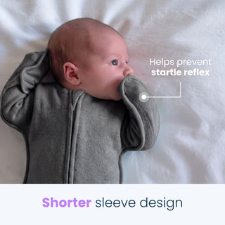 A baby wearing the HALO SwaddleStretch™ Newborn Swaddle TOG 0.5 with shorter sleeves rests on a white blanket. Text reads: Helps prevent startle reflex and Shorter sleeve design. The infant’s hands are positioned near their face, highlighting the hip-healthy sleep sack, designed for comfortable and secure swaddling.