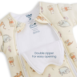 Close-up of the Disney® Winnie the Pooh Baby Merlin’s Magic Sleepsuit® 100% Cotton by HALO, featuring pastel-colored Winnie the Pooh characters on an ivory background. The sleepsuit showcases a double zipper design with a flap labeled “Double zipper For easy opening,” making it ideal for swaddle transitions or periods of sleep regression.