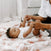 A baby in a white onesie lies on an aden + anais boutique cotton muslin dream blanket while holding hands with an adult wearing a white top. The scene is cozy and breathable, with soft lighting enhancing its peacefulness.