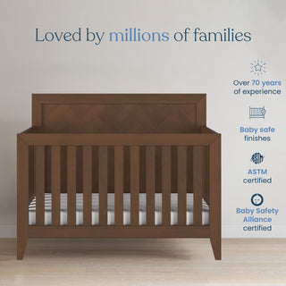The Child Craft Kieran 4-in-1 Convertible Crib in Toasted Chestnut features a striped mattress and stands against the wall. With 70+ years of expertise, it offers baby-safe finishes, ASTM and Baby Safety Alliance certifications.