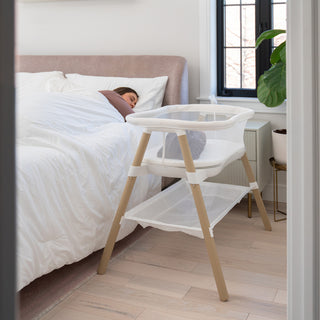 A woman sleeps in bed next to the HALO BassiNest® Essential Sleeper, a modern space-saving bassinet that's empty. The room has light wood floors, a plant, and a window with black frames.