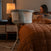 A woman sits on a bed with a rust quilt, holding a baby. Beside her is the white HALO® BassiNest® Swivel Sleeper 4.0 Signature by HALO. A lit lamp and nightstand are in the softly glowing background.