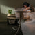 A woman leans over from her bed to reach into the HALO BassiNest® Swivel Sleeper 4.0 Air with Dreamweave Breathable Pad beside a nightstand holding a green potted plant, bathed in soft, warm light.
