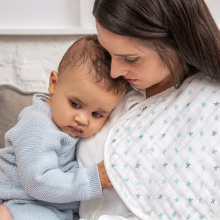 A woman comforts her baby, who’s in a light blue sweater. Draped over her shoulder is an aden + anais organic cotton snap bib with blue stars, part of their 3-pack set. Both share a calm, affectionate moment.
