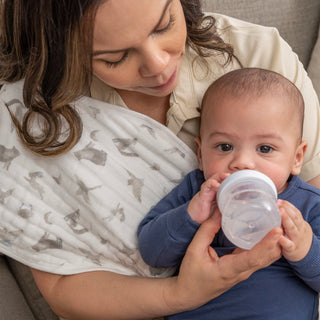 A woman bottle-feeds a baby in blue on the couch, using an aden + anais organic cotton muslin oversized washcloth as a burp cloth—an eco-friendly essential from the 3 pack.
