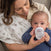 A woman bottle-feeds a baby in blue on the couch, using an aden + anais organic cotton muslin oversized washcloth as a burp cloth—an eco-friendly essential from the 3 pack.