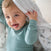A smiling baby in a teal long-sleeve shirt sticks out their tongue while holding an aden + anais organic cotton muslin swaddle blanket 2 pack above their head.
