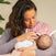 A woman with long dark hair cradles a baby, holding an aden + anais organic cotton muslin burpy bibs 2 pack over her shoulder. The soft background puts the focus on the cozy bib and tender moment.