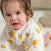 A baby with brown hair and blue eyes crawls on a soft surface, wearing a white outfit with yellow and orange fruit patterns and an aden + anais organic cotton muslin burpy bib from the 2 pack, looking curiously at the camera.