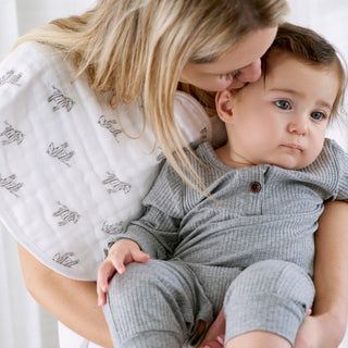 A woman kisses a baby in a gray outfit sitting on her lap, using the aden + anais Organic Baby Essentials Bundle. The zebra print burpy bib is featured, and both are indoors in soft natural light.