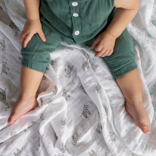 A baby sits on a white blanket featuring animal prints, wearing a green outfit. The blanket is the aden + anais organic cotton muslin burpy bibs 2 pack. The photo is taken from above, showing the baby's legs stretched out and one hand resting on their knee.