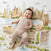 A smiling baby in a beige romper lies on an aden + anais organic cotton muslin dream blanket featuring a cityscape print, next to a wooden sign that says 6 months.